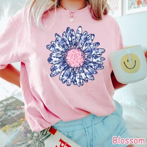 May include: A light pink t-shirt featuring a large daisy design. The daisy has blue and white patterned petals and a pink center. The word "Blossom" is printed in pink on the bottom right.