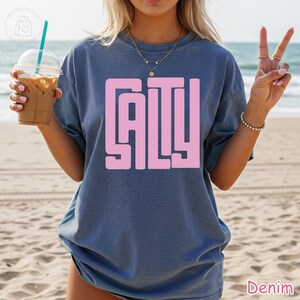 May include: Denim blue t-shirt with the word "SALTY" in a pink, block letter design. The shirt is a relaxed fit and has short sleeves. The image is taken on a beach.