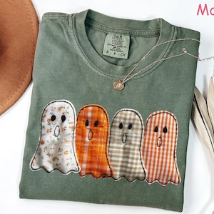 May include: Moss green t-shirt with four ghost appliqués in various patterns: floral, orange plaid, gray plaid, and orange and white plaid. The shirt has a crew neck and a small gold necklace. A brown hat and dried leaves are in the background.