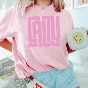 May include: A light pink sweatshirt with the word "SALTY" printed in large, pink, block letters. The person is holding a white mug with a yellow smiley face. The person is wearing light blue shorts.