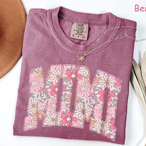 K&ouml;nnte beinhalten: Beerenfarbenes T-Shirt mit dem Wort "MOM" in einem floralen Design. Das Shirt hat einen Rundhalsausschnitt und ein kleines Etikett am Kragen. Eine goldene Halskette mit Anh&auml;nger ist &uuml;ber dem Shirt drapiert. Das Shirt ist Gr&ouml;&szlig;e S-P-CH.