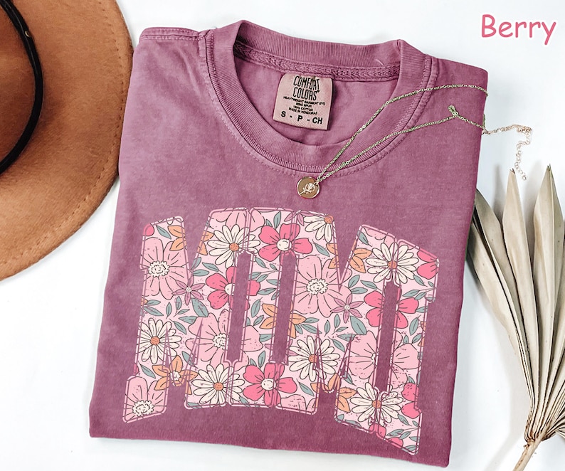 K&ouml;nnte beinhalten: Beerenfarbenes T-Shirt mit dem Wort "MOM" in einem floralen Design. Das Shirt hat einen Rundhalsausschnitt und ein kleines Etikett am Kragen. Eine goldene Halskette mit Anh&auml;nger ist &uuml;ber dem Shirt drapiert. Das Shirt ist Gr&ouml;&szlig;e S-P-CH.