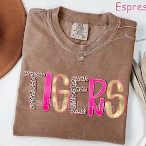 Peut inclure: T-shirt couleur expresso avec le mot "TIGERS" en rose et or. Les lettres "T" et "S" pr&eacute;sentent un motif l&eacute;opard. Un collier argent&eacute; avec un pendentif est pos&eacute; sur le t-shirt.