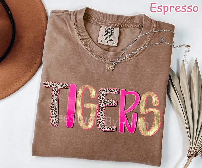 Peut inclure: T-shirt couleur expresso avec le mot "TIGERS" en rose et or. Les lettres "T" et "S" pr&eacute;sentent un motif l&eacute;opard. Un collier argent&eacute; avec un pendentif est pos&eacute; sur le t-shirt.