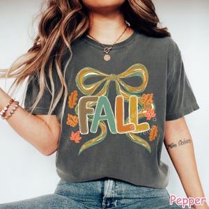 May include: Dark gray t-shirt with a colorful "FALL" graphic. The design features a large bow in shades of gold and teal, with the word "FALL" in brown, teal, and orange. Autumn leaves surround the text, creating a seasonal theme.