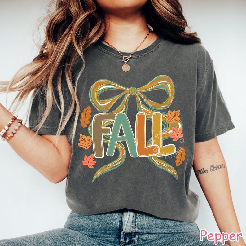 May include: Dark gray t-shirt with a colorful "FALL" graphic. The design features a large bow in shades of gold and teal, with the word "FALL" in brown, teal, and orange. Autumn leaves surround the text, creating a seasonal theme.