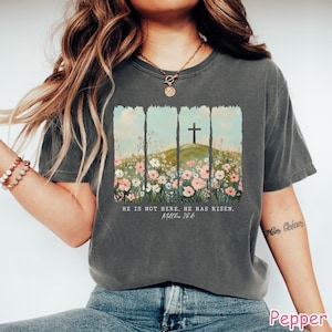 May include: A gray t-shirt with a floral design and a cross on a hill. The text "He is not here. He has risen. Matthew 28:6" is printed on the shirt.