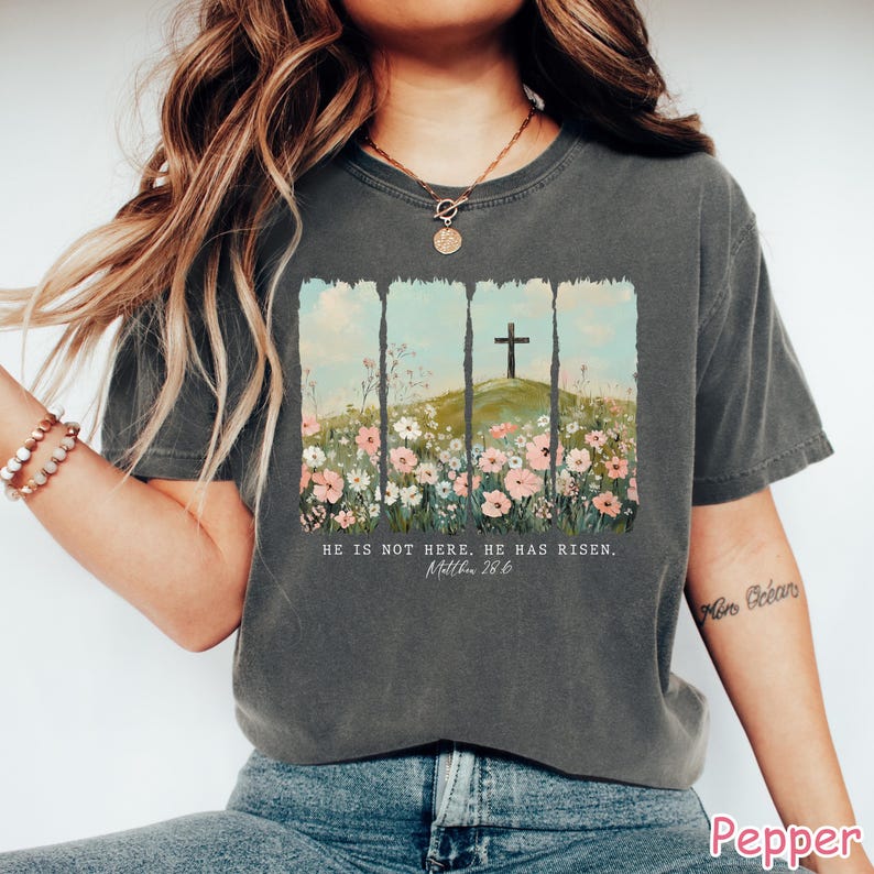 May include: A gray t-shirt with a floral design and a cross on a hill. The text "He is not here. He has risen. Matthew 28:6" is printed on the shirt.
