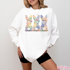 May include: White sweatshirt featuring three patchwork bunny designs in pastel colors. Each bunny has a bow and a floral accent. The sweatshirt is a long-sleeved, casual top, perfect for Easter or spring.