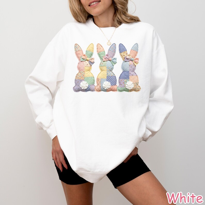 May include: White sweatshirt featuring three patchwork bunny designs in pastel colors. Each bunny has a bow and a floral accent. The sweatshirt is a long-sleeved, casual top, perfect for Easter or spring.