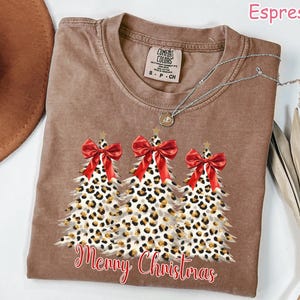 May include: Espresso-colored t-shirt featuring three Christmas trees with leopard print and red bows. The trees are adorned with gold glitter and topped with gold stars. The words "Merry Christmas" are written in red script below the trees.