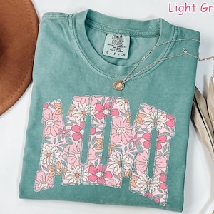 K&ouml;nnte beinhalten: Hellgr&uuml;nes T-Shirt mit dem Wort "MOM" in einem floralen Design. Die Buchstaben sind mit rosa, wei&szlig;en und orangefarbenen Blumen gef&uuml;llt. Eine silberne Halskette mit einem kleinen Anh&auml;nger liegt &uuml;ber dem Shirt.