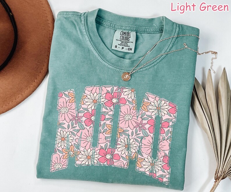 K&ouml;nnte beinhalten: Hellgr&uuml;nes T-Shirt mit dem Wort "MOM" in einem floralen Design. Die Buchstaben sind mit rosa, wei&szlig;en und orangefarbenen Blumen gef&uuml;llt. Eine silberne Halskette mit einem kleinen Anh&auml;nger liegt &uuml;ber dem Shirt.