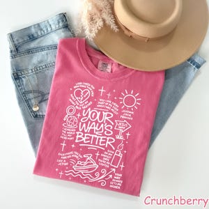 Op de afbeelding: Een roze t-shirt met witte tekst en afbeeldingen, waaronder de zin "YOUR WAY'S BETTER". Het shirt is opgevouwen en ligt op een blauwe spijkerbroek, een beige hoed en gedroogde bloemen. Het t-shirt is van het merk 'Comfort Colors'.