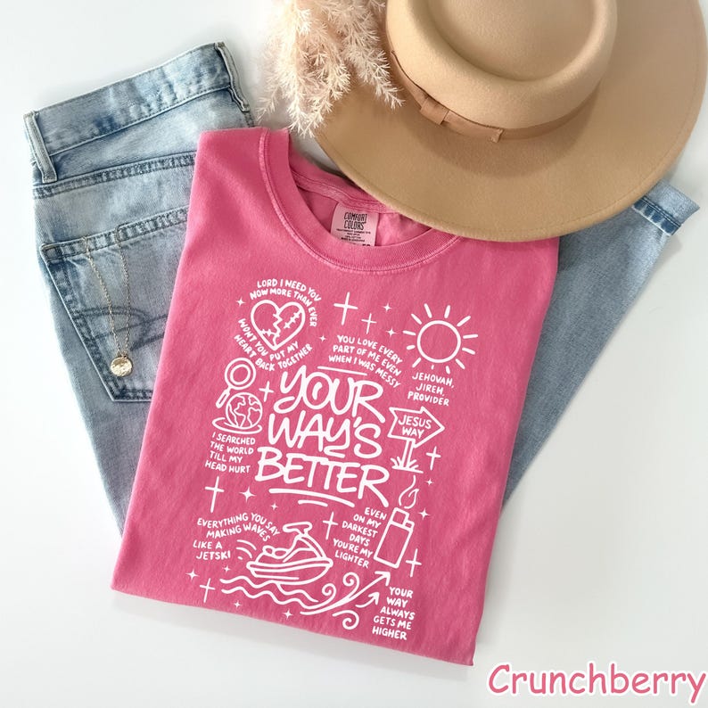 Op de afbeelding: Een roze t-shirt met witte tekst en afbeeldingen, waaronder de zin "YOUR WAY'S BETTER". Het shirt is opgevouwen en ligt op een blauwe spijkerbroek, een beige hoed en gedroogde bloemen. Het t-shirt is van het merk 'Comfort Colors'.