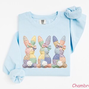 May include: Light blue sweatshirt featuring three appliqued bunny rabbits with patchwork designs in pastel colors. Each bunny has a bow and a white pom-pom tail. The word "Chambray" is printed in the bottom right corner.