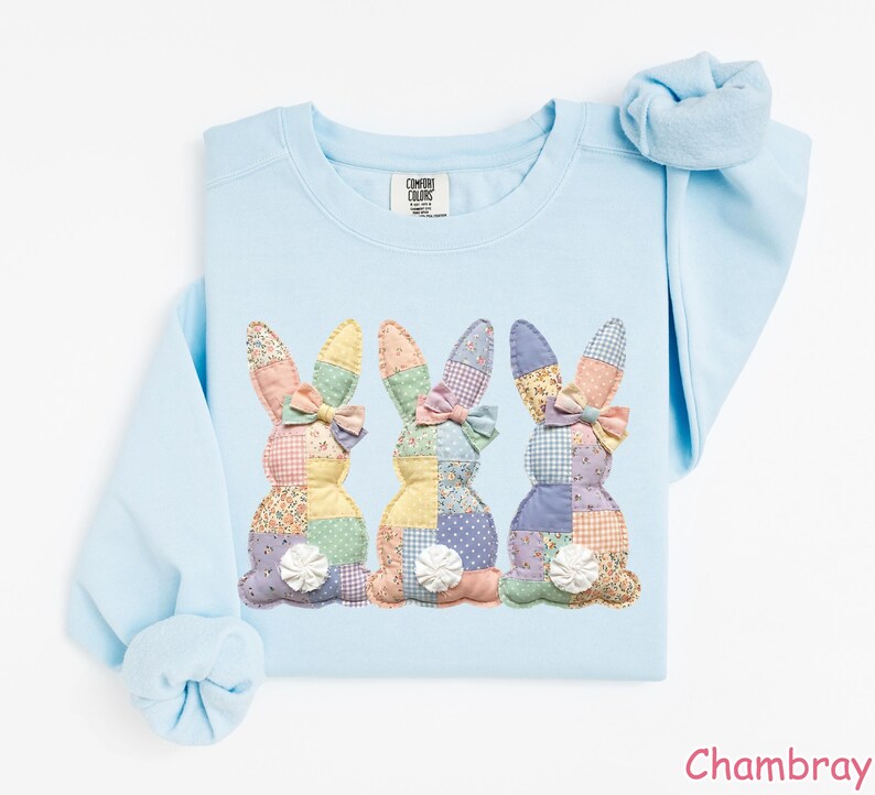 May include: Light blue sweatshirt featuring three appliqued bunny rabbits with patchwork designs in pastel colors. Each bunny has a bow and a white pom-pom tail. The word "Chambray" is printed in the bottom right corner.
