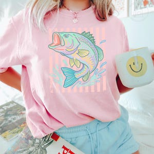 May include: A light pink short-sleeve t-shirt featuring a graphic of a jumping fish in shades of green, yellow, and blue. The fish is centred over a background of vertical light orange stripes. The shirt has a crew neckline and a relaxed fit.