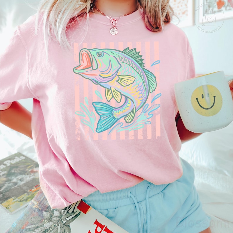 May include: A light pink short-sleeve t-shirt featuring a graphic of a jumping fish in shades of green, yellow, and blue. The fish is centred over a background of vertical light orange stripes. The shirt has a crew neckline and a relaxed fit.
