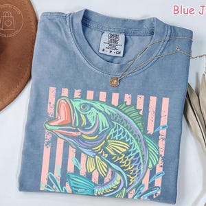 May include: A blue jean coloured t-shirt featuring a graphic print of a fish in shades of green, yellow, and purple. The design includes vertical light pink stripes behind the fish. The shirt has a crew neckline and a Comfort Colors brand label.