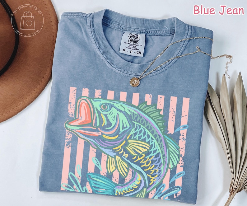 May include: A blue jean coloured t-shirt featuring a graphic print of a fish in shades of green, yellow, and purple. The design includes vertical light pink stripes behind the fish. The shirt has a crew neckline and a Comfort Colors brand label.