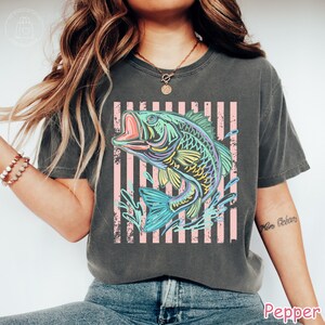 May include: A charcoal grey short-sleeve t-shirt featuring a graphic of a jumping fish in vibrant neon colours of blue, green, and yellow. The fish is set against a background of vertical light pink stripes with a distressed, weathered texture.