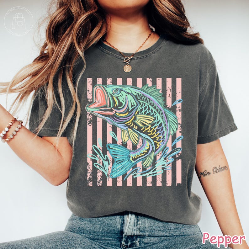 May include: A charcoal grey short-sleeve t-shirt featuring a graphic of a jumping fish in vibrant neon colours of blue, green, and yellow. The fish is set against a background of vertical light pink stripes with a distressed, weathered texture.
