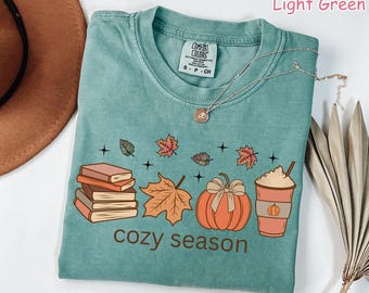 May include: Light green t-shirt with a graphic of books, leaves, a pumpkin, and a coffee cup. The text "cozy season" is printed in brown. A silver necklace is draped on the shirt.