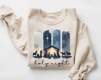 Könnte beinhalten: Sandfarbenes Sweatshirt mit einem Aquarelldesign einer Krippenszene in Marineblau und Gold. Das Design beinhaltet einen Stern, einen Stall und die Worte "holy night" in einer Schriftart.