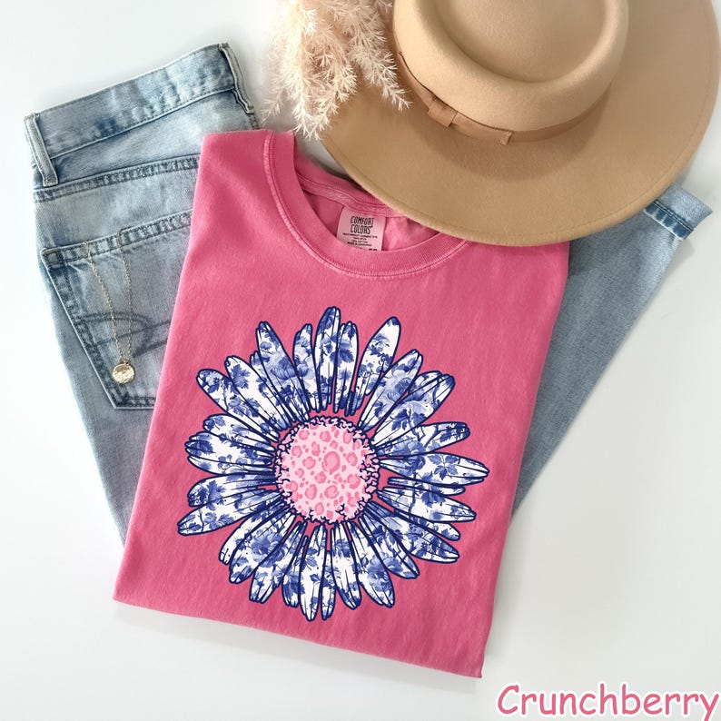 May include: A pink t-shirt with a blue and white floral design. The flower has a pink center with a leopard print pattern. The t-shirt is displayed on a white surface with a pair of blue jeans and a brown hat.