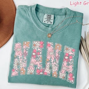 May include: Light green t-shirt with the word "MAMA" in large letters outlined with pink and orange flowers. The shirt is made of a soft, comfortable fabric and has a relaxed fit.