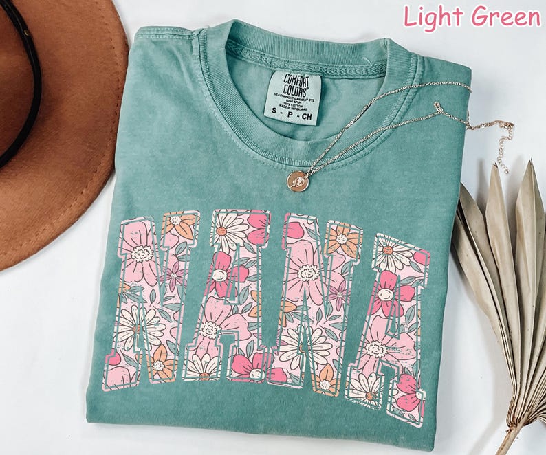 May include: Light green t-shirt with the word "MAMA" in large letters outlined with pink and orange flowers. The shirt is made of a soft, comfortable fabric and has a relaxed fit.