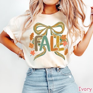 May include: Ivory-colored t-shirt with a colorful "FALL" graphic. The design features a large bow and autumn leaves in shades of green, orange, and brown. The word "FALL" is in a mix of colors. The shirt is paired with blue jeans.