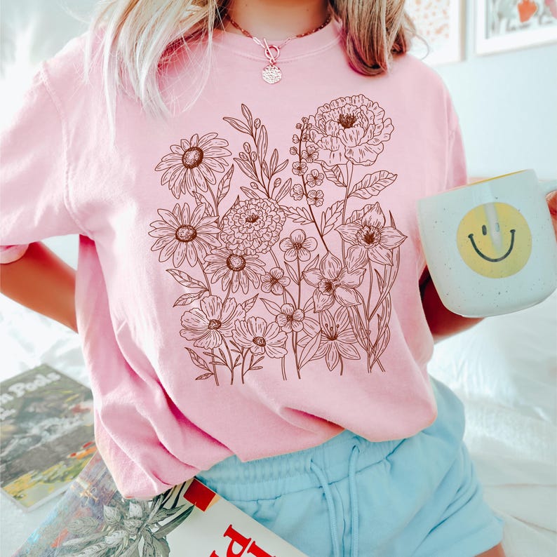 Puede incluir: Una sudadera rosa claro con un dise&ntilde;o floral marr&oacute;n. El dise&ntilde;o incluye varias flores y hojas. Se sostiene una taza blanca con una carita sonriente amarilla. La sudadera se combina con pantalones cortos azules.