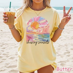 May include: Butter yellow t-shirt with a sunset graphic. The graphic features a colorful sun, water, and sky with the words "chasing sunsets" in black script. The shirt is a relaxed fit.