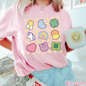 Peut inclure: Un t-shirt rose avec un motif &agrave; carreaux et diverses images de dessins anim&eacute;s color&eacute;s, notamment un arc-en-ciel, un fer &agrave; cheval, un tr&egrave;fle, une licorne, un c&oelig;ur et une &eacute;toile. Le t-shirt a un col rond et des manches courtes.