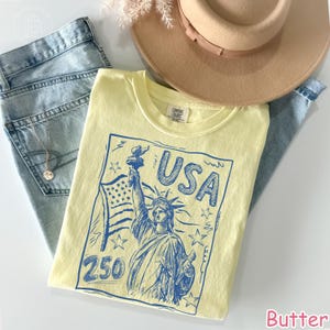 Puede incluir: Una camiseta de color amarillo p&aacute;lido con un estampado gr&aacute;fico azul de la Estatua de la Libertad, una bandera estadounidense, estrellas y el texto "USA 250". La camiseta se presenta sobre vaqueros azul claro, un sombrero beige y un collar.