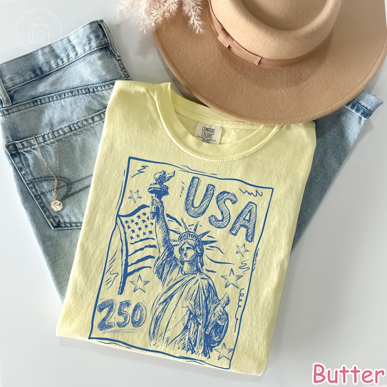 Puede incluir: Una camiseta de color amarillo p&aacute;lido con un estampado gr&aacute;fico azul de la Estatua de la Libertad, una bandera estadounidense, estrellas y el texto "USA 250". La camiseta se presenta sobre vaqueros azul claro, un sombrero beige y un collar.