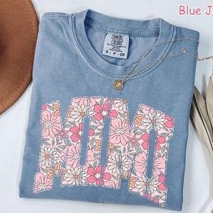 K&ouml;nnte beinhalten: Blaues Jeans-T-Shirt mit dem Wort "MOM" in einem floralen Design. Die Buchstaben sind mit rosa und wei&szlig;en Blumen gef&uuml;llt. Eine goldene Halskette mit Anh&auml;nger liegt &uuml;ber dem Shirt.