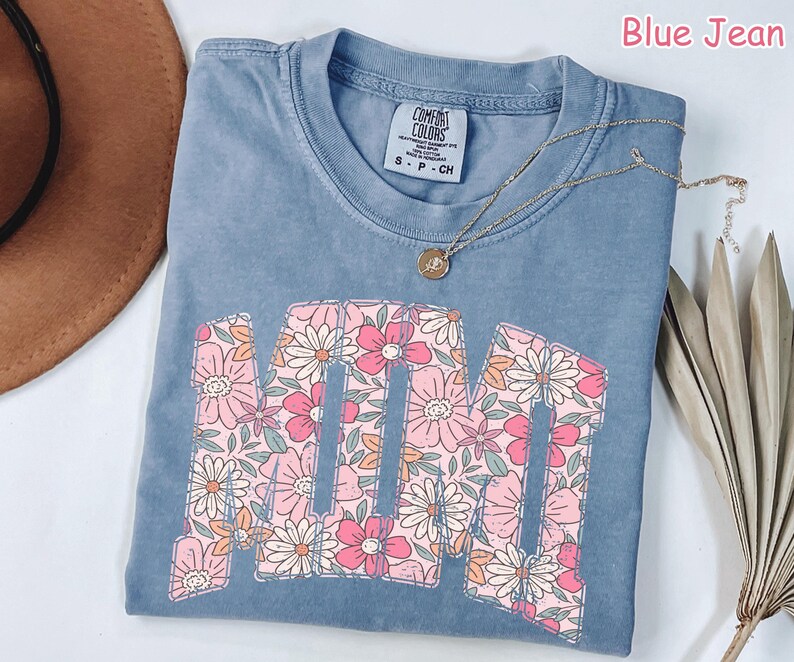 K&ouml;nnte beinhalten: Blaues Jeans-T-Shirt mit dem Wort "MOM" in einem floralen Design. Die Buchstaben sind mit rosa und wei&szlig;en Blumen gef&uuml;llt. Eine goldene Halskette mit Anh&auml;nger liegt &uuml;ber dem Shirt.