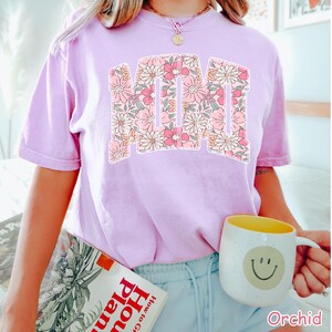 K&ouml;nnte beinhalten: Ein helllila T-Shirt mit dem Wort "MOM" in einem floralen Design. Das florale Design umfasst rosa, wei&szlig;e und pfirsichfarbene Blumen. Das Shirt ist ein kurz&auml;rmeliger Rundhalsstil. Das Wort "Orchid" ist unten rechts aufgedruckt.