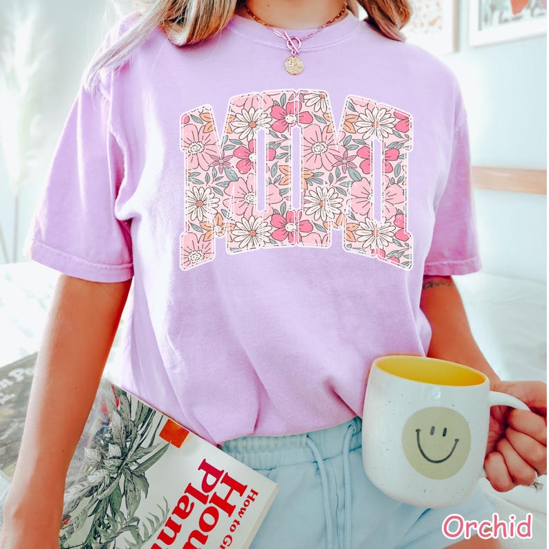 K&ouml;nnte beinhalten: Ein helllila T-Shirt mit dem Wort "MOM" in einem floralen Design. Das florale Design umfasst rosa, wei&szlig;e und pfirsichfarbene Blumen. Das Shirt ist ein kurz&auml;rmeliger Rundhalsstil. Das Wort "Orchid" ist unten rechts aufgedruckt.