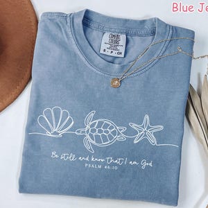 May include: Blue jean colored t-shirt with a white line drawing of a seashell, turtle, and starfish. The text "Be still and know that I am God, Psalm 46:10" is printed below the drawing. A gold necklace is draped across the shirt.