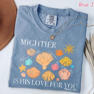 K&ouml;nnte beinhalten: Blaues Jeans-T-Shirt mit den Worten "MIGHTIER than the waves of the sea IS HIS LOVE FOR YOU" und Muschel-Grafiken. Eine goldene Halskette liegt &uuml;ber dem Shirt. Das Shirt ist von der Marke Comfort Colors.