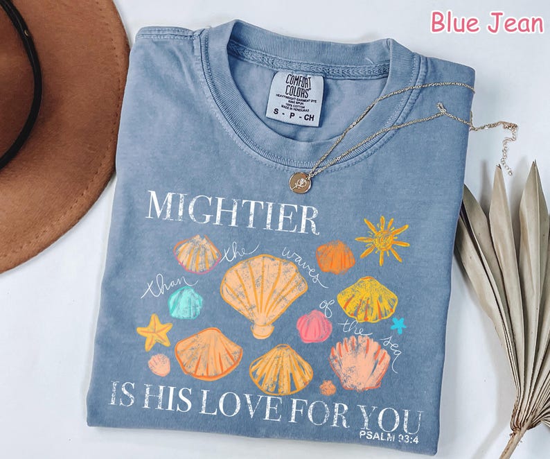 K&ouml;nnte beinhalten: Blaues Jeans-T-Shirt mit den Worten "MIGHTIER than the waves of the sea IS HIS LOVE FOR YOU" und Muschel-Grafiken. Eine goldene Halskette liegt &uuml;ber dem Shirt. Das Shirt ist von der Marke Comfort Colors.