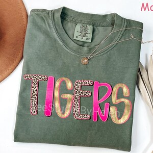 Peut inclure: T-shirt vert mousse avec le mot "TIGERS" dans un design superpos&eacute;. Les lettres ont un contour rose vif avec un remplissage dor&eacute;. Les lettres "T" et "S" ont un motif l&eacute;opard. Un collier dor&eacute; avec un pendentif est sur le t-shirt.
