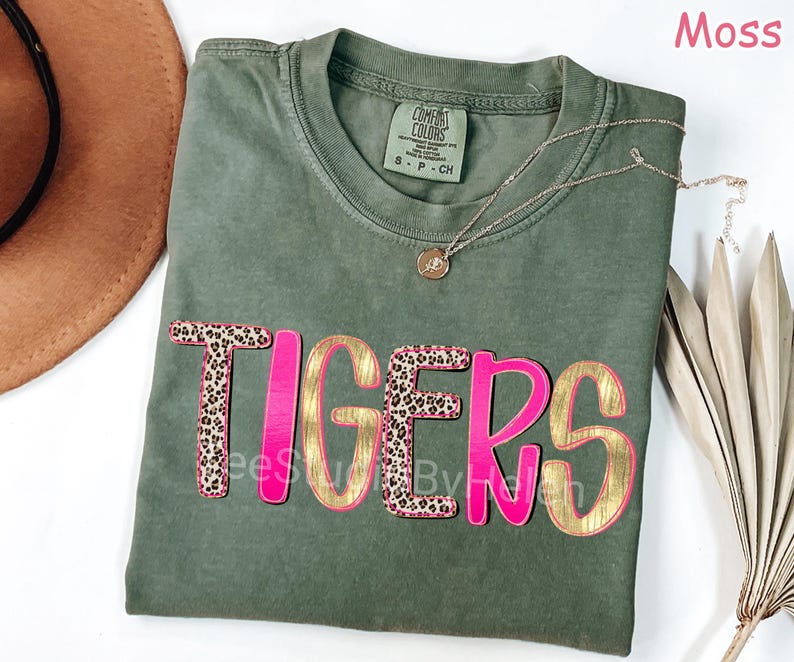 Peut inclure: T-shirt vert mousse avec le mot "TIGERS" dans un design superpos&eacute;. Les lettres ont un contour rose vif avec un remplissage dor&eacute;. Les lettres "T" et "S" ont un motif l&eacute;opard. Un collier dor&eacute; avec un pendentif est sur le t-shirt.