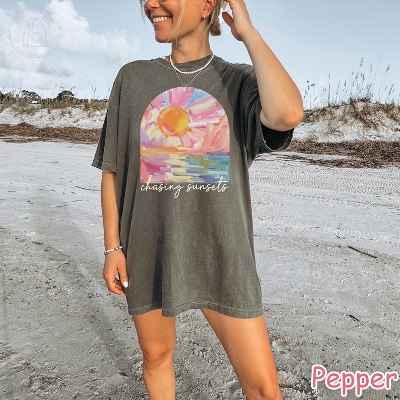 May include: A dark gray t-shirt with a colorful sunset graphic. The graphic features a sun, water, and the words "chasing sunsets". The shirt is a relaxed fit and is being worn on a beach.