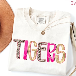 Peut inclure: T-shirt ivoire avec le mot "TIGERS" dans un design audacieux. Les lettres combinent un imprim&eacute; l&eacute;opard, de l'or et du rose, cr&eacute;ant un look vibrant. Un collier dor&eacute; et un chapeau marron sont &eacute;galement visibles.