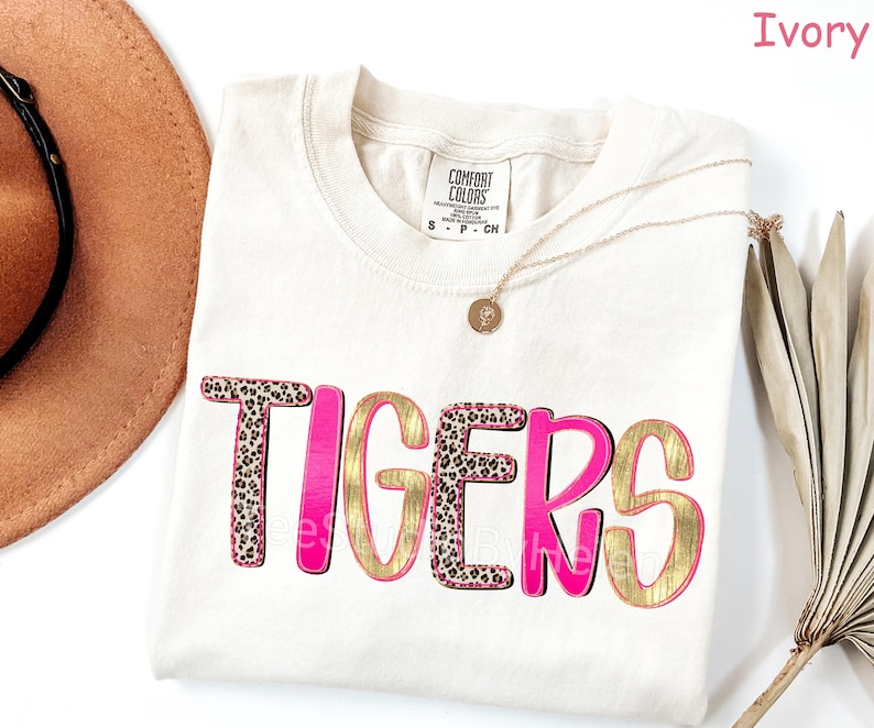 Peut inclure: T-shirt ivoire avec le mot "TIGERS" dans un design audacieux. Les lettres combinent un imprim&eacute; l&eacute;opard, de l'or et du rose, cr&eacute;ant un look vibrant. Un collier dor&eacute; et un chapeau marron sont &eacute;galement visibles.